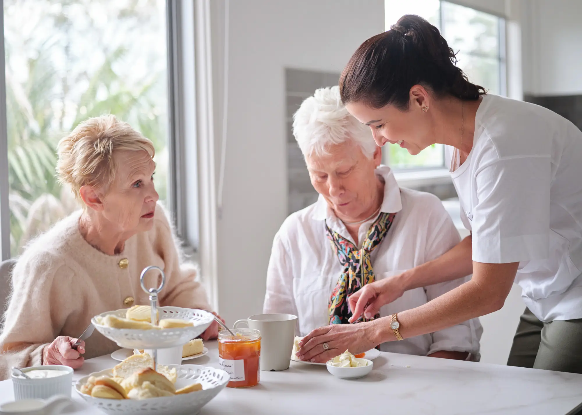 A home carer serving scones and tea to two older women in a bright, comfortable home.